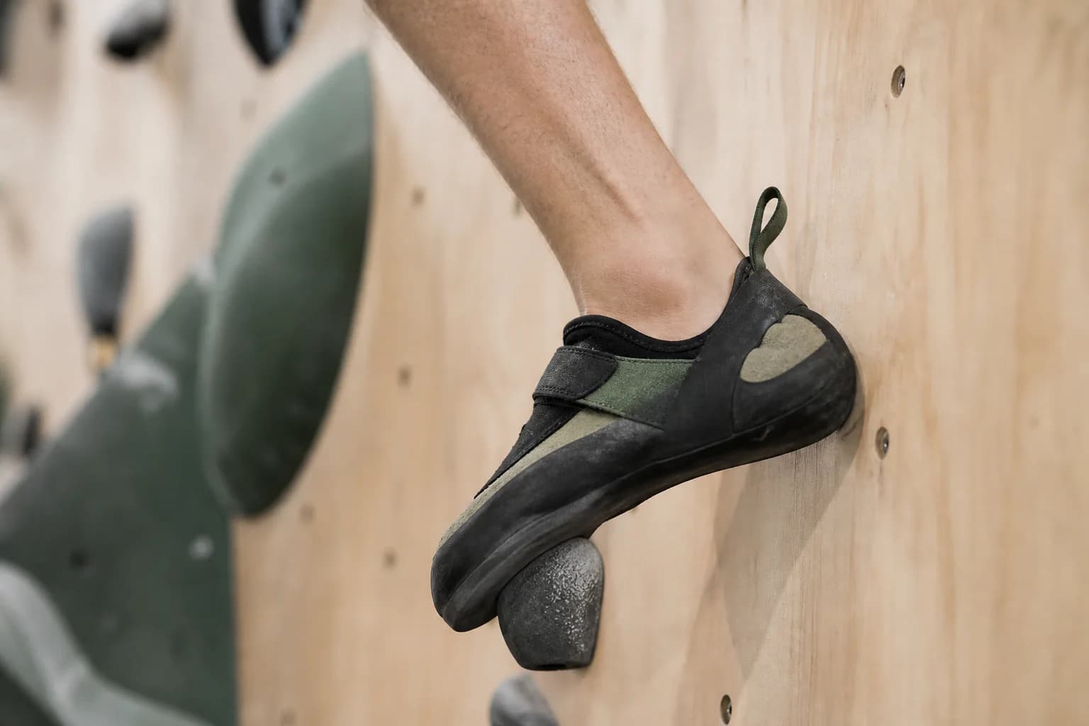 Close view of an olive climbing shoe weighting a small black foothold on a plywood climbing wall.