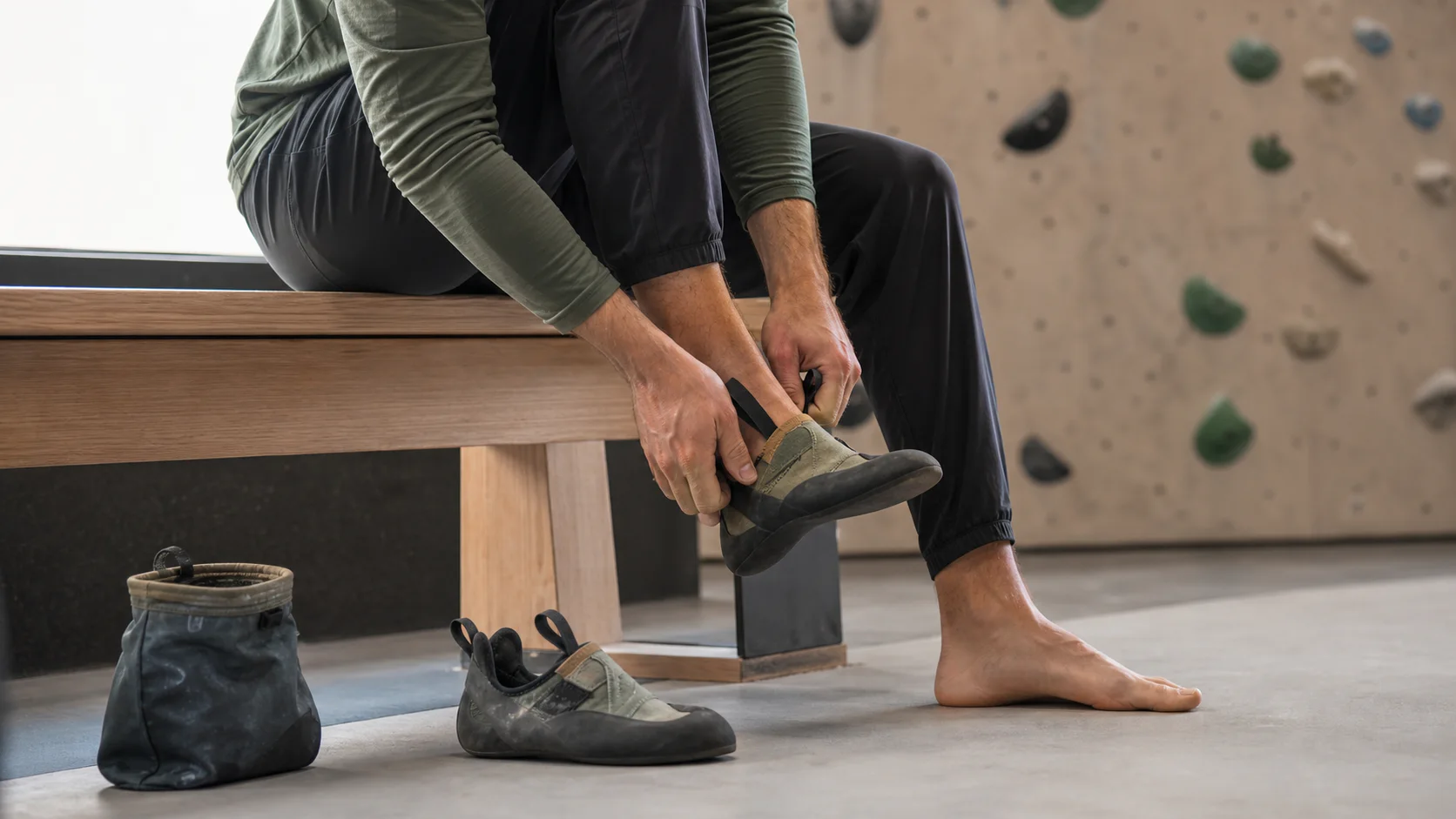 A climber seated on a wooden gym bench pulls on an olive climbing shoe while a matching shoe and chalk bag rest on the floor.
