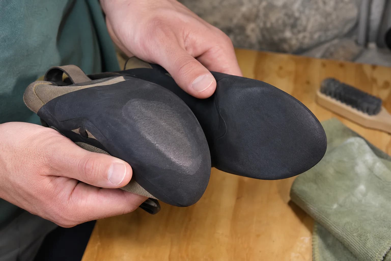 Hands hold two climbing-shoe soles together under a work light; one toe has a pale smooth wear patch and the other remains evenly black.